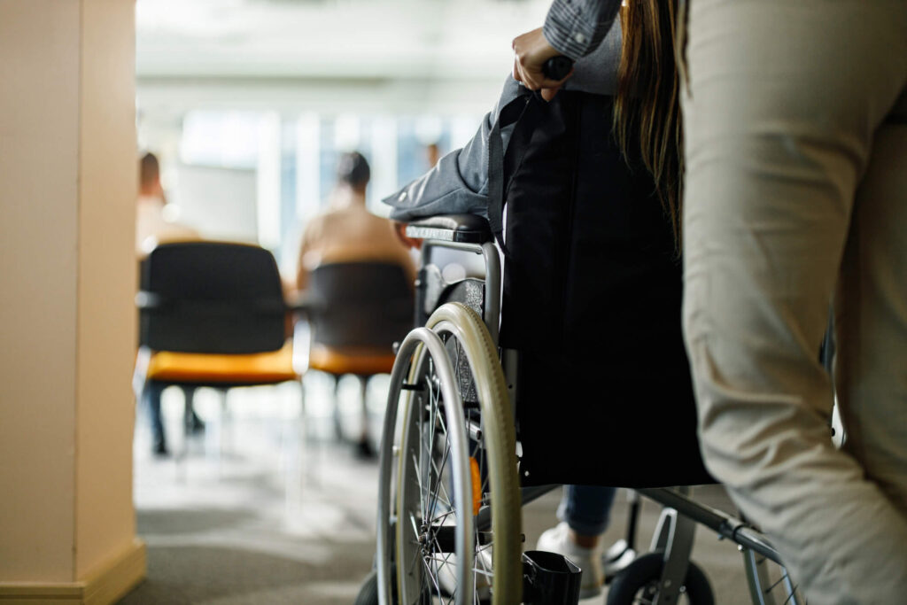 Image for the homepage to represent disability inclusion. It's an image of someone being pushed in a wheelchair to a meeting room.